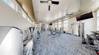 Located in the Southern Highlands community of Las Vegas, NV, this fitness center spans a single floor and is divided into two dedicated exercise rooms, offering a comprehensive workout environment. The space features an elegant yet functional design, with tray ceilings accented by neutral tones and modern Sputnik-style chandeliers that add a stylish touch to the athletic setting. Ceiling fans are installed throughout, ensuring comfortable airflow during workouts.

The cardio room is bright and inviting, lined with large windows that allow abundant natural light to fill the space and offer views of the surrounding neighborhood. It houses multiple True-brand elliptical machines and other cardio equipment, complemented by TRX suspension training straps mounted to a wall-mounted bar system. Large framed mirrors enhance the sense of openness.

The strength training room is equally well-appointed, featuring a full rack of dumbbells, adjustable workout benches, cable machines, and a variety of selectorized strength machines. A decorative geometric mirror panel serves as a striking design element, while wall-mounted flat-screen TVs provide entertainment. The textured stone accent walls and plush patterned carpet flooring tie both rooms together with a cohesive, upscale aesthetic throughout the facility.