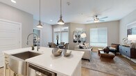 This thoughtfully designed single-floor residence in Phoenix, Arizona offers a seamless open-concept layout that connects the kitchen, dining, and living areas into one cohesive and functional space. The contemporary kitchen is a standout feature, showcasing gray shaker-style cabinetry with glass-front uppers, a white quartz waterfall island with bar seating, stainless steel appliances, a tile backsplash, and pendant lighting — all set atop warm hardwood-style flooring that extends throughout the main living areas. The dining area accommodates a full table and chairs, while the living room benefits from abundant natural light through large windows with white blinds and a ceiling fan overhead. Two well-appointed bedrooms offer plush carpeting, ceiling fans, generous windows with sheer drapery, and ample space for full bedroom suites. The primary bathroom features a dual-sink vanity with white countertops, taupe cabinetry, large-format tile flooring, and a separate water closet. The secondary bathroom includes a single vanity and a tub-shower combination. Neutral warm tones, recessed lighting, and clean architectural lines give the entire residence a polished, modern ambiance throughout.