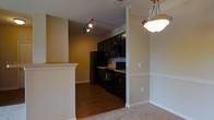 This contemporary apartment showcases a thoughtfully designed open floor plan, seamlessly integrating the kitchen and living spaces. The kitchen, visible through a partial wall opening, boasts sleek black cabinetry and appliances, creating a striking contrast against light-colored countertops. Track lighting above ensures ample task illumination. 

The living area, distinguished by plush carpeting, offers a cozy atmosphere. A prominent feature is the elegant dome light fixture, adding a touch of sophistication to the space. The transition between rooms is marked by a change in flooring, with the kitchen featuring durable wood-look flooring.

Neutral wall colors throughout promote a sense of spaciousness and provide a versatile backdrop for personalization. Crown molding and baseboards add refined architectural details. The partially visible entryway suggests additional living space beyond, hinting at a well-planned layout designed for modern living.