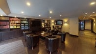 This well-appointed party room is designed for upscale entertaining, seamlessly combining a fully equipped kitchen with a comfortable and stylish lounge area. The kitchen is outfitted with dark cabinetry, stainless steel appliances including a double wall oven, gas cooktop, and refrigerator, complemented by a curved island with seating — perfect for casual dining or mingling. Rich hardwood flooring flows throughout the space, lending warmth and continuity between areas. A wide hallway with recessed and wall-mounted accent lighting connects the various zones, creating an inviting sense of arrival. The lounge area is furnished with dark leather club chairs and low wooden coffee tables, arranged to encourage conversation and relaxation. Built-in bookshelves filled with books flank a large flat-screen television, adding a refined, club-like atmosphere. Floor-to-ceiling sliding glass doors along the lounge wall flood the space with natural light and offer sweeping views of the surrounding wooded landscape. Recessed lighting throughout ensures the space is well-lit while maintaining an intimate ambiance. Together, these elements create a versatile, elegant environment suited for a wide range of social events.