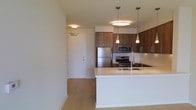 This single-floor residence offers a thoughtfully designed open-concept layout that seamlessly blends functionality with contemporary style. The kitchen serves as a central highlight, showcasing rich, warm-toned wood cabinetry that provides ample storage alongside a full suite of stainless steel appliances, including a gas range, built-in microwave, and a French door refrigerator. A generous kitchen island with a clean white countertop anchors the space, offering additional workspace and a double undermount sink with a modern arc faucet. Stylish pendant lights hang above the island, complementing the flush-mount ceiling fixtures that provide bright, even illumination throughout. Light hardwood-style flooring runs throughout the space, adding warmth and continuity to the open floor plan. The entry foyer is well-defined, featuring a solid door with secure hardware that transitions naturally into the main living area. The overall ambiance is clean, bright, and inviting, with a neutral color palette of whites and warm wood tones that creates a modern yet approachable aesthetic, making the space both practical and visually appealing.