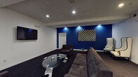 Located in the heart of Los Angeles, CA, the Regency Lounge is a thoughtfully designed single-floor space that blends bold aesthetics with comfortable functionality. The lounge is immediately distinguished by its striking cobalt blue accent wall, which serves as a dramatic focal point against the surrounding white walls and dark carpeted flooring.

The main seating area is furnished with a large tufted gray sectional sofa and matching armchairs, all arranged around a sleek, sculptural glass coffee table with chrome legs. A wall-mounted flat-screen television anchors one side of the room, making the space well-suited for both casual gatherings and media viewing.

A pair of tall, high-back tufted white wingback chairs with dark wood frames occupies a more intimate corner of the lounge, accompanied by a small round glass side table — offering a refined, conversation-friendly seating nook. Recessed lighting throughout the ceiling provides warm, focused illumination, complementing the lounge's upscale ambiance.

A dedicated service area features a modern coffee and beverage station atop a curved glass console table, adding a hospitality-forward element to the space. A decorative geometric patterned wall hanging adds texture and visual interest to the blue accent wall. The overall atmosphere is polished and contemporary, designed to provide guests with a premium, comfortable lounge experience.