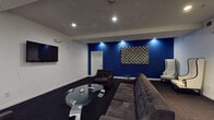 Located in the heart of Los Angeles, CA, the Regency Lounge is a thoughtfully designed single-floor space that blends bold aesthetics with comfortable functionality. The lounge is immediately distinguished by its striking cobalt blue accent wall, which serves as a dramatic focal point against the surrounding white walls and dark carpeted flooring.

The main seating area is furnished with a large tufted gray sectional sofa and matching armchairs, all arranged around a sleek, sculptural glass coffee table with chrome legs. A wall-mounted flat-screen television anchors one side of the room, making the space well-suited for both casual gatherings and media viewing.

A pair of tall, high-back tufted white wingback chairs with dark wood frames occupies a more intimate corner of the lounge, accompanied by a small round glass side table — offering a refined, conversation-friendly seating nook. Recessed lighting throughout the ceiling provides warm, focused illumination, complementing the lounge's upscale ambiance.

A dedicated service area features a modern coffee and beverage station atop a curved glass console table, adding a hospitality-forward element to the space. A decorative geometric patterned wall hanging adds texture and visual interest to the blue accent wall. The overall atmosphere is polished and contemporary, designed to provide guests with a premium, comfortable lounge experience.