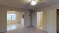 Located at 215 Whitehall Ln in Jacksonville, North Carolina, this two-story home offers a practical and comfortable layout suited for everyday living. The main level features a welcoming open-concept design where the living area flows naturally toward the entryway and kitchen. The living room is finished with plush carpeting and is equipped with a ceiling fan with light fixture, providing both comfort and airflow. A half-wall partition separates the kitchen area, which transitions into tile flooring and benefits from natural light provided by a window near the front door. The entryway is bright and accessible, featuring a classic paneled front door. Adjacent to the living area, a door leads to a closet space, offering convenient storage. The neutral color palette throughout the main level — with warm beige walls and light-toned finishes — creates a clean, move-in-ready atmosphere. The textured ceiling adds a traditional character to the space. With two floors, the home provides separation between living and private spaces, making it well-suited for a variety of lifestyles. The overall design is functional and straightforward, with attention to practical details throughout.