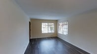 This single-floor space presents a clean, modern aesthetic defined by a thoughtful combination of simplicity and functionality. The main living area immediately draws attention with its rich, dark hardwood-style flooring, which contrasts elegantly against the soft, neutral-toned walls and crisp white baseboards and ceiling. The room is generously illuminated by natural light streaming through multiple large windows fitted with horizontal blinds, offering both brightness and privacy control. The windows frame views of the surrounding exterior greenery, adding a refreshing connection to the outdoors. The open layout of the living space provides a versatile foundation, allowing for a variety of furniture arrangements to suit different needs. The smooth, white ceiling contributes to an airy and open ambiance throughout. A doorway visible along one wall suggests seamless connectivity to additional areas of the unit. Overall, the space strikes a balance between practicality and comfort, with its well-maintained finishes, abundant natural light, and neutral palette creating a welcoming environment suitable for a range of uses.