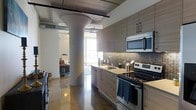 This single-floor space showcases a compelling blend of industrial and contemporary design. The open-concept layout is anchored by a striking sculptural column that elegantly transitions from a wide base to a tapered form, serving as both a structural element and a defining architectural feature. The kitchen is thoughtfully appointed with flat-front cabinetry in a warm taupe wood finish, stainless steel appliances including an electric range, over-the-range microwave, and a mosaic tile backsplash that adds texture and visual depth. Light-toned countertops provide ample workspace, while the seamless flow into the living area enhances the sense of openness. Polished concrete floors run throughout, reflecting natural light and contributing to the loft's industrial character. Exposed ductwork and conduit along the ceiling reinforce the raw, urban aesthetic. Large windows at the far end of the space flood the interior with abundant natural light, creating a bright and inviting atmosphere. The living area, visible beyond the column, complements the kitchen's modern palette. Overall, the space offers a cohesive, stylish environment that balances functional design with distinctive architectural personality.