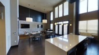This single-floor communal space showcases a sleek, contemporary design defined by soaring double-height ceilings that create an expansive and airy atmosphere. The dark hardwood flooring grounds the space, providing a rich contrast against the neutral tones of the walls and furnishings. Large windows dressed with horizontal blinds line the upper portion of one wall, allowing abundant natural light to filter throughout the room. A prominent accent wall clad in dark horizontal shiplap paneling serves as a dramatic focal point, flanked by a wall-mounted flat-screen television and a complementary stone-textured tile panel, adding depth and visual interest. Multiple seating clusters, each featuring round tables and upholstered chairs, are thoughtfully arranged across the space to encourage conversation and collaboration. Overhead, a series of large cylindrical pendant lights hang at varying heights, providing warm, ambient illumination. A sleek white countertop surface in the foreground hints at an integrated serving or reception area. Double glass entry doors with sidelights connect the interior to the outdoors, enhancing the sense of openness. Overall, the space balances modern elegance with functional comfort.