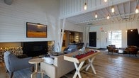This inviting single-floor space masterfully blends rustic charm with modern design. The interior is defined by dramatic vaulted ceilings clad in white shiplap paneling, supported by painted structural beams that add both character and architectural interest. Warm Edison bulb pendant lights hang at varying heights, casting a cozy, ambient glow throughout the open-plan layout.

The main living area centers around a sleek, modern fireplace flanked by a neatly stacked wood pile, creating a natural focal point for relaxation and gathering. A flat-screen television is mounted above, adding a contemporary touch. Comfortable seating, including plush sofas and tufted accent chairs, is arranged to encourage conversation and leisure.

A distinctive communal table with X-shaped white legs and a rich wood top takes center stage, adorned with vintage-style blue glass accents. The warm, wide-plank hardwood flooring runs throughout, grounding the space with natural texture and warmth.

Beyond the main seating area, a secondary lounge or workspace is visible, separated by a classic sliding barn door, reinforcing the space's rustic-modern aesthetic. The open layout, soaring ceiling height, and thoughtful mix of materials create an atmosphere that is both spacious and welcoming.