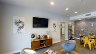 This well-appointed single-floor space offers a thoughtfully designed interior that blends modern aesthetics with functional comfort. The main lounge area serves as the focal point, featuring a wall-mounted flat-screen television positioned above a mid-century modern media console adorned with curated decorative accents, including a globe, potted greenery, and eclectic accessories that add character and warmth to the space.

Recessed lighting illuminates the room from above, casting a clean, even light across the light gray walls and patterned carpeted flooring. A pair of contemporary swivel accent chairs in a muted blue-gray tone provide comfortable seating, complemented by additional soft furnishings that contribute to the inviting atmosphere.

Abstract wall art flanks the television, lending an artistic touch to the overall design. A sleek open shelving unit along the adjacent wall displays an assortment of decorative objects, further enhancing the curated feel of the interior.

A hallway visible from the lounge leads to additional rooms, suggesting a well-connected floor plan. The space maintains a cohesive, modern design language throughout, with clean lines, neutral tones, and carefully selected furnishings that create a polished and welcoming environment suitable for both relaxation and social interaction.