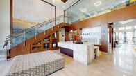 The Park 3400 Clubhouse in Davie, Florida is a thoughtfully designed community facility that blends contemporary architecture with upscale interior finishes. The entrance foyer welcomes visitors with polished large-format tile flooring, warm wood-paneled walls, and sculptural decor, leading directly to the branded reception area. The reception desk is a standout feature, crafted from richly veined marble and set against a backdrop of wood paneling displaying the PARC3400 logo. A striking staircase with glass railings and a large-scale abstract painting anchors the entryway, adding an artistic focal point to the space. The grand main lounge boasts soaring double-height ceilings and expansive floor-to-ceiling windows that flood the interior with natural light and offer views of the lush, palm-lined outdoor surroundings. Comfortable leather seating arrangements are anchored by vibrant teal and gold area rugs beneath elegant pendant lighting. Additional lounge areas throughout the clubhouse feature contemporary sectional sofas, colorful original artwork, and teal accent ottomans, creating inviting spaces for socializing or relaxation. The overall ambiance is modern and energetic, with a cohesive palette of teal, orange, and warm wood tones lending a lively yet polished character to the entire facility.