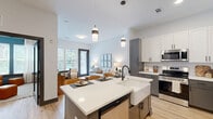 This stylish single-floor residence features a cohesive modern design with light wood flooring, crisp white walls, and dark-accented doors and hardware that create a refined, contemporary aesthetic throughout. The open-concept kitchen is a standout, showcasing two-tone cabinetry in white and gray, quartz countertops, stainless steel appliances, and a large center island with a farmhouse sink and pendant lighting — ideal for both cooking and casual dining. The kitchen flows seamlessly into the living room, which is bathed in natural light from oversized windows and a glass door that opens to an outdoor view of the surrounding cityscape. The bedroom offers a comfortable, well-proportioned retreat with generous natural light and access to a walk-in closet. Two full bathrooms are finished with granite countertops, gray cabinetry, black hardware, and wood-look flooring, maintaining the home's consistent modern sensibility. A welcoming hallway connects the living areas to the private spaces, adding to the intuitive flow of the floor plan. Throughout, the design balances practicality with elevated finishes, resulting in a polished and livable space.