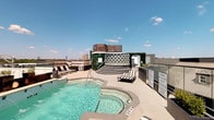 This rooftop pool deck offers a well-appointed outdoor retreat with a thoughtfully designed layout that balances relaxation and recreation. The centerpiece is a sparkling swimming pool accompanied by an adjacent hot tub, both finished with clean, contemporary surroundings. Striped lounge chairs are arranged poolside for sunbathing, while a raised platform area with stylish wood-paneled accents and lush vertical greenery provides a distinct focal point and gathering space.

The deck extends beyond the pool area to include open-air terraces finished in large-format light stone tiles. These spaces are furnished with compact seating arrangements, ideal for socializing or taking in the surroundings. Tall sculptural planters with greenery add touches of natural life throughout.

A seamless glass perimeter railing preserves unobstructed panoramic views of the city skyline and surrounding tree canopy, making the outdoor experience both visually expansive and connected to the urban environment. The open-sky setting ensures abundant natural light throughout the day.

Additional amenities include outdoor dining and communal table seating in a more sheltered section of the rooftop, offering practical space for casual gatherings. The overall design blends modern aesthetic sensibilities with functional outdoor living, creating a rooftop destination that is both inviting and visually striking.