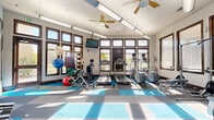 Located at 3310 S Kenton St in Denver, CO, this fitness center offers a single, thoughtfully organized exercise room designed to support a comprehensive workout routine. The space is bathed in natural light, thanks to an expansive array of large, dark-framed windows that line multiple walls and extend toward the ceiling, providing open outdoor views and an energizing atmosphere. Ceiling fans and industrial-style overhead lighting contribute to a comfortable and well-ventilated environment.

The flooring features a distinctive blue and gray tile pattern that zones the space visually while providing durable, functional surfaces suitable for varied exercise activities. Cardio equipment is well-represented, with multiple treadmills, an elliptical, a stationary bike, and a recumbent cycle positioned to take advantage of the natural light. The strength training area includes a squat/power rack with a flat bench, a multi-station cable machine, and additional resistance equipment. A dedicated free weights section offers a rack of dumbbells alongside stability balls and medicine balls for functional fitness training.

Wall-mounted flat-screen televisions are positioned throughout the room for entertainment during workouts. A large mirror along one wall enhances the sense of space and allows users to monitor their form. Hand sanitizer stations are also present, reflecting a commitment to cleanliness and user well-being.