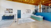 Located at 25 W Seventh St in Richmond, VA, this single-floor commercial space offers a generously scaled, open interior defined by a compelling blend of industrial and contemporary design elements. The architecture is immediately distinguished by soaring wood-beamed ceilings with tongue-and-groove planking, exposed brick walls, and polished concrete floors that extend throughout the entire footprint.

Floor-to-ceiling steel-framed windows line the street-facing facade, flooding the interior with abundant natural light while offering clear views of the surrounding neighborhood. A dramatic double-sided stone fireplace anchors the central portion of the space, serving as a visual and functional dividing feature between distinct seating areas. Rope-wrapped structural columns add an organic, textural contrast to the refined finishes.

A bold teal staircase, adorned with a curated gallery wall of framed artwork, leads to an upper level and serves as a strong focal point. Near the entry, a teal fluted reception desk sits beneath vaulted white walls, providing a polished welcome area. Pendant lighting with globe and chandelier fixtures enhances the warm ambiance. The combination of lounge seating, communal dining areas, and strategic furnishing arrangements makes the space well-suited for a variety of commercial or hospitality uses.