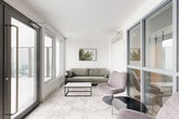 Located at 150 Rue King in Montréal, this mezzanine-level lounge is a compact yet well-appointed space that blends modern design with a warm, inviting atmosphere. The room is defined by its striking marble-effect porcelain tile flooring, which runs throughout and lends an air of elegance and visual spaciousness to the interior.

Large windows along the exterior wall flood the space with natural light, while a set of full-height French doors provides direct access to an outdoor balcony, offering views of the surrounding urban environment. A wall-mounted mini-split climate control unit ensures year-round comfort.

The layout is thoughtfully arranged into two distinct seating areas. One area features a sage green upholstered sofa paired with a minimalist black-framed coffee table, anchored by botanical wall art that reinforces the space's nature-inspired aesthetic. The second area offers a pair of wooden sling chairs with geometric-patterned upholstery, arranged around a wire-frame side table beside a towering Bird of Paradise plant, which adds a lush, organic element to the crisp white surroundings.

Bright white walls and a clean white ceiling enhance the sense of openness, while the carefully curated furnishings create a cohesive, contemporary feel. This mezzanine lounge serves as a refined and functional communal or transitional space within the building.
