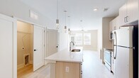 Located at 98 Northwood Blvd in Pittsboro, NC, this well-designed one-bedroom, one-bathroom residence offers a clean, modern aesthetic throughout its thoughtfully arranged single-floor layout. The open-concept kitchen and living area serves as the heart of the home, anchored by a generous kitchen island with a white quartz countertop, an undermount sink, and light wood cabinetry. Decorative pendant lighting hangs above the island, complementing the stainless steel appliances and light hardwood flooring that flows seamlessly into the living area. The living room is bright and airy, featuring a ceiling fan, large windows with horizontal blinds, and a glass-panel door that opens directly onto a private balcony — ideal for enjoying the outdoors. The bedroom offers soft carpeting, warm neutral walls, and ample natural light from multiple windows. The bathroom features a wide vanity with white quartz countertops, light wood cabinetry with matte black hardware, and a large framed mirror, conveying a sense of clean, modern style. Throughout the space, a consistent palette of warm whites and soft beige tones, combined with natural light and modern finishes, creates a bright and welcoming ambiance.