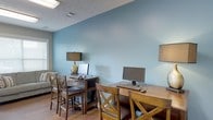 This single-floor space functions as a dedicated computer lounge or business center, designed to offer a functional and comfortable environment for work or personal use. The room is finished with distinctly cool-toned, steel-blue walls that lend the space a calm, focused atmosphere, complemented by warm hardwood flooring that adds a grounding contrast to the cool palette overhead.

Two wooden workstations are arranged along the walls, each paired with matching X-back wooden chairs that echo a classic design sensibility. One desk hosts a laptop setup accompanied by a decorative table lamp with a gold base and a black shade, providing localized task lighting. The second workstation is equipped with a full desktop monitor, keyboard, and mouse, offering a more traditional computing setup. An additional lamp is partially visible, contributing to the overall warm ambient lighting alongside the overhead fluorescent fixture.

The textured ceiling adds a subtle architectural detail to the room. Ventilation is provided via visible ceiling vents, ensuring comfort during use. The space is compact yet efficiently arranged to accommodate multiple users simultaneously, making it well-suited as a shared business center or study area.