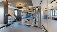 This expansive fitness center is designed with a contemporary aesthetic, offering a comprehensive range of workout zones across a single level. The space is defined by its striped, multi-tone rubberized flooring, which visually separates functional areas while providing durable, shock-absorbing surfaces throughout. Oversized circular pendant lights hang from coffered ceilings, casting warm, focused illumination across the equipment while contributing to the upscale ambiance.

The cardio zone features treadmills, stair climbers, and elliptical machines positioned near floor-to-ceiling windows that invite abundant natural light and outward views of the surrounding area. Full-length mirrors line multiple walls, visually expanding the space and supporting form-conscious training.

A dedicated strength training section houses cable machines, adjustable benches, and a multi-station cable system by InLight Fitness, alongside a rack of color-coded dumbbells and stability balls for varied workouts. A glass-partitioned functional training area contains resistance bands, a freestanding pad station, and rubber flooring suited for dynamic movement.

Artistic touches, including a bold athletic mural on the glass partition entry, reinforce the motivational atmosphere. The overall design balances high-end finishes with practical functionality, creating a polished and inviting environment for a full-spectrum fitness experience.