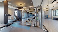 This expansive fitness center is designed with a contemporary aesthetic, offering a comprehensive range of workout zones across a single level. The space is defined by its striped, multi-tone rubberized flooring, which visually separates functional areas while providing durable, shock-absorbing surfaces throughout. Oversized circular pendant lights hang from coffered ceilings, casting warm, focused illumination across the equipment while contributing to the upscale ambiance.

The cardio zone features treadmills, stair climbers, and elliptical machines positioned near floor-to-ceiling windows that invite abundant natural light and outward views of the surrounding area. Full-length mirrors line multiple walls, visually expanding the space and supporting form-conscious training.

A dedicated strength training section houses cable machines, adjustable benches, and a multi-station cable system by InLight Fitness, alongside a rack of color-coded dumbbells and stability balls for varied workouts. A glass-partitioned functional training area contains resistance bands, a freestanding pad station, and rubber flooring suited for dynamic movement.

Artistic touches, including a bold athletic mural on the glass partition entry, reinforce the motivational atmosphere. The overall design balances high-end finishes with practical functionality, creating a polished and inviting environment for a full-spectrum fitness experience.