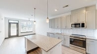 Located at 1300 Front Ave in Columbus, GA, this modern one-bedroom, one-bathroom unit offers a thoughtfully designed open-concept layout. The combined kitchen and living area serves as the heart of the home, anchored by a large granite-topped kitchen island with built-in shelving beneath. The kitchen is well-appointed with gray shaker-style cabinetry, white subway tile backsplash, granite countertops, and a full suite of stainless steel appliances including a refrigerator, range, microwave, and dishwasher. Stylish pendant lighting hangs above the island, adding a refined touch. Light wood-style flooring flows throughout the main living areas, enhancing the bright, airy ambiance. The living area benefits from large windows fitted with plantation-style blinds that invite generous natural light, along with a ceiling fan for comfort. The bedroom is carpeted and well-lit by a broad window overlooking the outdoors, and features a ceiling fan and direct access through gray panel doors to both the bathroom and a walk-in closet with wire shelving systems. The bathroom is clean and modern, offering a granite vanity, gray cabinetry, a framed mirror, and a glass-enclosed shower with tile surround.