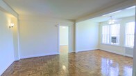 This single-floor space is defined by its classic architectural details and well-maintained interior. The main living area is immediately notable for its gleaming parquet hardwood flooring, arranged in a traditional basket-weave pattern that adds warmth and timeless character to the space. Neutral, cream-toned walls provide a clean backdrop that enhances the sense of brightness throughout.

Natural light enters through a large window fitted with vertical blinds, illuminating the open and airy atmosphere of the room. The smooth, white ceiling complements the light walls, contributing to an overall sense of spaciousness.

A doorway connects the main living area to an adjoining room, which continues the same parquet flooring, ensuring a cohesive aesthetic throughout. The passage is framed by clean white trim and molding, consistent with the space's understated, classic style. White baseboards run along the perimeter of the rooms, adding a polished finishing touch.

The layout is straightforward and functional, making it well-suited for a variety of uses. The space presents a neutral, move-in-ready canvas that offers both comfort and versatility.