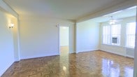 This single-floor space is defined by its classic architectural details and well-maintained interior. The main living area is immediately notable for its gleaming parquet hardwood flooring, arranged in a traditional basket-weave pattern that adds warmth and timeless character to the space. Neutral, cream-toned walls provide a clean backdrop that enhances the sense of brightness throughout.

Natural light enters through a large window fitted with vertical blinds, illuminating the open and airy atmosphere of the room. The smooth, white ceiling complements the light walls, contributing to an overall sense of spaciousness.

A doorway connects the main living area to an adjoining room, which continues the same parquet flooring, ensuring a cohesive aesthetic throughout. The passage is framed by clean white trim and molding, consistent with the space's understated, classic style. White baseboards run along the perimeter of the rooms, adding a polished finishing touch.

The layout is straightforward and functional, making it well-suited for a variety of uses. The space presents a neutral, move-in-ready canvas that offers both comfort and versatility.
