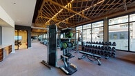 This fitness center is a thoughtfully designed, single-floor facility that combines industrial aesthetics with a comprehensive workout environment. The space is defined by its standout architectural feature: an exposed wood-lattice ceiling with integrated linear lighting that spans the main exercise floor, creating a warm, visually dynamic atmosphere. Floor-to-ceiling windows line one side of the facility, flooding the interior with abundant natural light and offering views of the surrounding building exterior.

The main workout floor is generously proportioned and accommodates a wide array of equipment, including a row of treadmills positioned along the windowed wall, ellipticals, stationary bikes, rowing machines, a stair climber, and a cable machine. A dedicated dumbbell rack with adjustable benches provides ample space for strength training, while a wall-mounted flat-screen television and full-length mirrors enhance the cardio area, which also features exposed brick accent walls.

A separate spin studio is enclosed with large interior windows overlooking the main floor, outfitted with professional spinning bikes, step platforms, stability balls, and mirrored walls with warm-framed accents and sleek linear overhead lighting.

Near the entrance, a convenient amenity station features built-in wood cubbies, a compact refrigerator, a water fountain, and a motivational neon "Sweat the Details" sign, rounding out the facility's welcoming and functional design.