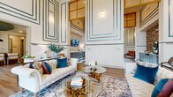 This thoughtfully designed single-floor space offers a generous footprint that seamlessly connects two primary areas: a formal living room and a combined kitchen-lounge zone. Dramatic double-height ceilings are a defining architectural feature, amplifying the sense of openness and grandeur throughout. Walls are adorned with teal-accented geometric molding panels that extend from floor to ceiling, creating a refined, art-deco-inspired aesthetic.

The living area is anchored by large floor-to-ceiling windows that flood the space with natural light and offer views of the surrounding outdoor area. Wide-plank hardwood-style flooring adds warmth and continuity. A foyer-style entry features a rustic console table and grid-patterned drapes framing tall windows.

The kitchen-lounge area transitions dramatically with a bold black-and-white checkerboard marble tile floor. Sage-toned cabinetry with white countertops lines one wall alongside a textured brick accent wall, while a wood-toned bar counter with bar seating provides a functional gathering point. A statement cage-style chandelier illuminates the space overhead. Together, both areas deliver a cohesive blend of classic elegance and contemporary comfort.