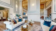 This thoughtfully designed single-floor space offers a generous footprint that seamlessly connects two primary areas: a formal living room and a combined kitchen-lounge zone. Dramatic double-height ceilings are a defining architectural feature, amplifying the sense of openness and grandeur throughout. Walls are adorned with teal-accented geometric molding panels that extend from floor to ceiling, creating a refined, art-deco-inspired aesthetic.

The living area is anchored by large floor-to-ceiling windows that flood the space with natural light and offer views of the surrounding outdoor area. Wide-plank hardwood-style flooring adds warmth and continuity. A foyer-style entry features a rustic console table and grid-patterned drapes framing tall windows.

The kitchen-lounge area transitions dramatically with a bold black-and-white checkerboard marble tile floor. Sage-toned cabinetry with white countertops lines one wall alongside a textured brick accent wall, while a wood-toned bar counter with bar seating provides a functional gathering point. A statement cage-style chandelier illuminates the space overhead. Together, both areas deliver a cohesive blend of classic elegance and contemporary comfort.