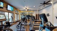 This fitness center is a thoughtfully designed, single-floor facility built to support a comprehensive workout experience. The space is generously proportioned, offering room for both cardiovascular and strength training activities without feeling cramped. A row of True-brand treadmills and cardio machines lines one side, while a variety of weight and resistance training equipment occupies the central and opposite areas. Large windows along one wall flood the room with natural light, creating a bright and energizing atmosphere, while multiple glass-paneled doors provide easy access and enhance the open feel. Ceiling fans are evenly distributed throughout, ensuring consistent airflow and comfort during workouts. Wall-mounted flat-screen TVs are positioned for visibility from multiple workout stations, adding an entertainment element to training sessions. A floor-to-ceiling mirror spans one wall, visually expanding the space and allowing users to monitor their form. Recessed lighting supplements the natural light, keeping the facility well-illuminated at all hours. The flooring is durable and fitness-appropriate, designed to withstand heavy equipment and high foot traffic. Overall, the space presents a clean, functional, and welcoming environment suitable for a wide range of fitness routines.