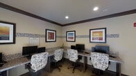 This business center is a thoughtfully designed single-floor space built to accommodate guests or residents seeking a productive work environment. The room features multiple computer workstations arranged along built-in granite-topped counters that line the walls, providing ample workspace for users. Each station is equipped with a desktop computer and monitor, paired with comfortable floral-patterned ergonomic chairs that add a decorative touch to the functional setting. The walls are finished in warm neutral tones and accented by a distinctive mosaic tile border that runs horizontally at mid-wall height, adding texture and visual interest to the space. A series of bold, colorful framed artworks are displayed above the tile border, contributing to the room's polished and welcoming ambiance. Recessed lighting in the ceiling ensures the space is well-lit throughout. The flooring features neutral-toned carpet, complementing the overall warm color palette. A posted rules board near the entrance provides users with clear guidelines for the space. The overall design strikes a balance between a professional work environment and a comfortable, inviting atmosphere.