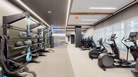 Located at 1100 4th St SW in Washington, DC, this professionally designed fitness center offers a generously sized, single-floor layout thoughtfully organized into distinct functional zones. The space features a contemporary aesthetic defined by a neutral palette of white, gray, and charcoal, accented by warm wood ceiling beams and striking blue LED accent lighting throughout.

The main exercise floor is divided into clearly delineated areas. A cardio zone houses a row of treadmills, ellipticals, stair climbers, and stationary bikes aligned along floor-to-ceiling frosted glass windows that invite natural light and exterior views. The strength training area features power racks, barbells, weight plates, adjustable benches, and a comprehensive dumbbell rack. A dedicated functional training zone includes rowing machines, resistance bands, stability balls, and open stretching space.

A separate studio room with hardwood floors, bold geometric wallcovering, blue LED accent lighting, and a large wall mirror provides space for group classes or individual training. Linear LED ceiling fixtures illuminate the entire facility evenly, complemented by recessed downlights. Storage shelving, a small service counter, and a private recovery room with a dedicated entrance further enhance the facility's comprehensive amenities.