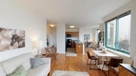 This two-bedroom, two-bathroom residence at 1 Alexander North in Yonkers, NY offers a well-designed single-floor layout with a clean, contemporary aesthetic. The open-concept living area is anchored by warm hardwood flooring and benefits from generous natural light streaming through large corner windows, which frame views of the surrounding streetscape. The living space flows seamlessly into the dining area, creating a comfortable and functional shared zone ideal for everyday living and entertaining.

The kitchen is equipped with warm-toned wood cabinetry, dark granite countertops, black appliances including a gas range, refrigerator, and built-in microwave, and a peninsula with bar seating that opens directly to the main living area. Tiled flooring grounds the kitchen space and transitions naturally to the hardwood beyond.

Both bedrooms are carpeted and offer a calm, neutral atmosphere. Each is well-proportioned, featuring windows with natural light and closet space. One bedroom includes an open reach-in closet, while the other provides a quiet, restful retreat.

The two bathrooms maintain a consistent modern style, featuring dark wood vanities with light countertops, mirrored medicine cabinets, and tile flooring. One bathroom includes a shower-tub combination, while the other is configured as a more compact full bath.