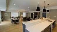 This single-floor space showcases a thoughtfully designed open-concept layout that seamlessly integrates the kitchen, dining, and living areas into one cohesive environment. The kitchen features a generous island topped with crisp white quartz countertops and accented by navy-blue cabinetry, anchored overhead by striking black pendant lights that add a contemporary focal point. The light hardwood flooring extends throughout, creating visual continuity and warmth across the entire space. The dining area comfortably accommodates a large table with upholstered seating, positioned near bright windows dressed with white horizontal blinds that allow natural light to filter in. The living area centers around a classic fireplace flanked by wall sconces, with a wall-mounted television above, creating an inviting gathering space. Deep charcoal sofas and woven accent chairs contribute a layered, textural quality to the seating arrangement. Crown molding, recessed lighting throughout the ceiling, and a neutral taupe-and-gray color palette lend the space a refined, polished ambiance. Overall, the design balances modern sophistication with everyday livability.
