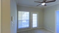 This single-floor space offers a clean, functional layout designed for comfortable everyday living. The main living area is a standout feature, bathed in natural light streaming through large windows dressed with horizontal blinds. A glass-panel exterior door sits alongside the windows, providing direct outdoor access while further enhancing the brightness of the room. The soft, neutral wall tones complement the carpeted flooring, creating a cohesive and understated aesthetic throughout. Crown molding trims the ceiling, adding a refined architectural detail that elevates the overall finish of the space. A ceiling fan with an integrated light fixture is centrally mounted, offering both practical airflow and supplemental lighting. The room's layout feels open and unobstructed, making it versatile for a variety of furniture arrangements. The combination of natural light, outdoor connectivity, and classic finishing touches gives the space a welcoming and well-maintained character. Overall, this space presents a straightforward yet thoughtfully finished environment suited for those seeking comfort and practicality in a single-level setting.