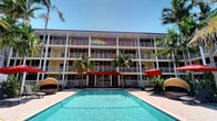 This multi-story building showcases a classic Florida resort-style architectural design, characterized by clean horizontal lines, a light-toned exterior, and expansive white-railed balconies that run the full width of each floor. The building's façade is punctuated by large windows and sliding glass doors that allow natural light to flow into each unit while offering direct access to private outdoor balconies with open views of the grounds below.

At the heart of the property lies a generously sized outdoor swimming pool with crystal-clear turquoise water, framed by a spacious travertine-style pool deck. The deck is furnished with lounge chairs and canopied pod seating, providing comfortable spots for relaxation. Bold red market umbrellas add a vibrant pop of color to the outdoor living area, complementing the warm tones of the building's exterior.

Tall, mature palm trees are strategically placed throughout the property, lending a lush, tropical character to the overall setting and providing natural shade. The landscaping enhances the resort-like atmosphere that defines the space. A covered entrance with a distinctive red awning serves as the focal point of the building's ground-level façade, anchoring the design with a welcoming and polished entryway. The entire setting conveys a relaxed yet well-appointed tropical lifestyle.