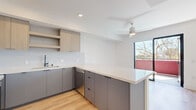 This thoughtfully designed single-floor residence offers three bedrooms, two bathrooms, a kitchen, and a closet, all finished in a clean, contemporary style. Light hardwood flooring runs throughout, complementing the bright white walls and creating a cohesive, modern aesthetic.

The kitchen is a standout feature, showcasing sleek gray flat-front cabinetry paired with light wood upper cabinets and open shelving. A white quartz island provides generous prep and gathering space, while a matte black faucet, stainless steel dishwasher, and a ceiling fan overhead add both function and style. Large black-framed windows invite abundant natural light and outdoor views into the kitchen and living areas.

A well-proportioned hallway connects the bedrooms, fitted with louvered closet doors and clean white walls. The primary bedroom is notably spacious, offering room to configure a full suite of furnishings. Additional bedrooms are bright and airy, with oversized black-framed windows that frame exterior views and flood the rooms with sunlight.

The two bathrooms are finished with large-format gray tile. One features a soaking tub with a tiled surround, while the other offers a walk-in shower with a window and matte black fixtures. Both include wood-toned vanities with white countertops and mirrors. The overall ambiance is modern, clean, and move-in ready.