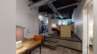 This single-floor loft showcases a compelling blend of industrial and contemporary design elements. The ceiling immediately draws the eye, featuring exposed metal ductwork, dark-stained wood beams, and structural columns that speak to the space's raw architectural character. The open-concept layout is defined by a notable split-level design, where a few steps separate the main living area from an elevated dining zone, adding visual depth and dimension to the floor plan.

The living area features a large sectional sofa positioned against a whitewashed exposed brick wall, which serves as a striking textural backdrop. Natural light filters through roller-shaded windows, and a sculptural round wood coffee table anchors the seating arrangement. The kitchen is efficiently designed with stainless steel appliances, including a full-size refrigerator and electric range, complemented by a slate-blue glass tile backsplash and dark countertops.

Two bedrooms offer calm retreats from the loft's industrial aesthetic, with smooth walls and understated décor. The bathroom is clean and modern, featuring a dark-cabinetry vanity with a white countertop, tiled flooring, and a walk-in shower with a curtain enclosure.