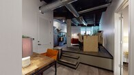 This single-floor loft showcases a compelling blend of industrial and contemporary design elements. The ceiling immediately draws the eye, featuring exposed metal ductwork, dark-stained wood beams, and structural columns that speak to the space's raw architectural character. The open-concept layout is defined by a notable split-level design, where a few steps separate the main living area from an elevated dining zone, adding visual depth and dimension to the floor plan.

The living area features a large sectional sofa positioned against a whitewashed exposed brick wall, which serves as a striking textural backdrop. Natural light filters through roller-shaded windows, and a sculptural round wood coffee table anchors the seating arrangement. The kitchen is efficiently designed with stainless steel appliances, including a full-size refrigerator and electric range, complemented by a slate-blue glass tile backsplash and dark countertops.

Two bedrooms offer calm retreats from the loft's industrial aesthetic, with smooth walls and understated décor. The bathroom is clean and modern, featuring a dark-cabinetry vanity with a white countertop, tiled flooring, and a walk-in shower with a curtain enclosure.