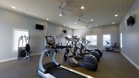 This single-floor fitness center is thoughtfully designed to support a comprehensive workout experience. The space is outfitted with a lineup of quality cardio equipment, including Precor elliptical machines and treadmills, arranged to allow comfortable movement throughout the room. Additional strength and resistance machines are positioned toward the back, offering a well-rounded selection of training options for users of all fitness levels.

The room features neutral gray walls and a low-pile carpet flooring that provides cushioning underfoot. Recessed lighting is distributed evenly across the ceiling, ensuring the space is well-lit at all times, while multiple ceiling fans promote air circulation and comfort during workouts. Large windows along one wall allow natural light to fill the room, contributing to an open and energizing atmosphere.

Wall-mounted flat-screen televisions are positioned at strategic angles, allowing exercisers to stay entertained during cardio sessions. The overall layout is clean, functional, and uncluttered, making it easy to navigate between equipment. The space strikes a balance between practicality and comfort, providing a welcoming environment for regular fitness routines.