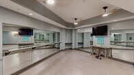 Located in Columbia, SC, this dedicated yoga and fitness studio is a thoughtfully designed single-room space that offers a clean, functional environment for exercise and wellness activities. The studio features a standout wall of floor-to-ceiling mirrors paired with horizontal ballet barres, providing practitioners with clear visual feedback during yoga, barre, dance, or general fitness sessions. The open floor plan is finished with neutral-toned tile flooring, offering ample room for individual or group workouts. A wall-mounted flat-screen television is positioned for easy viewing from anywhere in the room, supporting instructor-led or self-guided routines. The tray ceiling adds architectural dimension to the space, while recessed lighting and pendant fixtures provide even, well-distributed illumination throughout. Gray-painted walls give the studio a modern, focused aesthetic. A rustic wooden utility table offers surface space for equipment or accessories, and colorful artwork accents the walls, adding a touch of personality to the otherwise streamlined environment. Small free weights and exercise balls visible in the space reflect its multi-use functionality. The studio is well-suited for yoga, barre, stretching, and light fitness training, making it a versatile and practical space for a range of wellness disciplines.