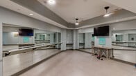 Located in Columbia, SC, this dedicated yoga and fitness studio is a thoughtfully designed single-room space that offers a clean, functional environment for exercise and wellness activities. The studio features a standout wall of floor-to-ceiling mirrors paired with horizontal ballet barres, providing practitioners with clear visual feedback during yoga, barre, dance, or general fitness sessions. The open floor plan is finished with neutral-toned tile flooring, offering ample room for individual or group workouts. A wall-mounted flat-screen television is positioned for easy viewing from anywhere in the room, supporting instructor-led or self-guided routines. The tray ceiling adds architectural dimension to the space, while recessed lighting and pendant fixtures provide even, well-distributed illumination throughout. Gray-painted walls give the studio a modern, focused aesthetic. A rustic wooden utility table offers surface space for equipment or accessories, and colorful artwork accents the walls, adding a touch of personality to the otherwise streamlined environment. Small free weights and exercise balls visible in the space reflect its multi-use functionality. The studio is well-suited for yoga, barre, stretching, and light fitness training, making it a versatile and practical space for a range of wellness disciplines.