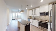 Located at 2801–2897 Bryan St in Dallas, TX 75204, this single-floor residence sits within the vibrant Uptown neighborhood and showcases a thoughtfully designed, contemporary interior. The open-concept kitchen serves as the centerpiece of the living area, featuring white shaker cabinetry with glass-front upper panels, dark granite countertops, a herringbone tile backsplash, and a full suite of stainless steel appliances including a range, microwave, and dishwasher. A generous kitchen island with matching dark granite and a built-in beverage cooler provides additional prep and gathering space, while polished pendant lights overhead add a refined industrial touch. Rich hardwood-style flooring extends through the main living areas, creating a warm contrast to the bright white walls and recessed lighting throughout. Large windows and a glass door at the end of the living area invite abundant natural light and offer outdoor views. The bedroom features soft carpeting and clean, neutral walls ready for personalization. The bathroom is appointed with a dual-sink vanity topped with dark granite, polished chrome fixtures, a wall-mounted mirror, and distinctive patterned tile flooring that adds a decorative flair. Overall, the space balances modern finishes with a clean, functional layout suited for everyday living.