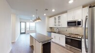 Located at 2801–2897 Bryan St in Dallas, TX 75204, this single-floor residence sits within the vibrant Uptown neighborhood and showcases a thoughtfully designed, contemporary interior. The open-concept kitchen serves as the centerpiece of the living area, featuring white shaker cabinetry with glass-front upper panels, dark granite countertops, a herringbone tile backsplash, and a full suite of stainless steel appliances including a range, microwave, and dishwasher. A generous kitchen island with matching dark granite and a built-in beverage cooler provides additional prep and gathering space, while polished pendant lights overhead add a refined industrial touch. Rich hardwood-style flooring extends through the main living areas, creating a warm contrast to the bright white walls and recessed lighting throughout. Large windows and a glass door at the end of the living area invite abundant natural light and offer outdoor views. The bedroom features soft carpeting and clean, neutral walls ready for personalization. The bathroom is appointed with a dual-sink vanity topped with dark granite, polished chrome fixtures, a wall-mounted mirror, and distinctive patterned tile flooring that adds a decorative flair. Overall, the space balances modern finishes with a clean, functional layout suited for everyday living.