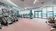 Located at 125 NE 32nd St in Miami's vibrant Edgewater neighborhood, this contemporary fitness center offers a generously sized, single-floor layout designed to accommodate a full spectrum of workout disciplines. The space is bathed in natural light, thanks to expansive floor-to-ceiling glass walls that frame views of the surrounding outdoor areas, including what appears to be a resort-style pool deck lined with lush greenery.

The main cardio zone is impressively equipped with a row of treadmills, stair climbers, and elliptical trainers, all positioned along the glass perimeter to allow exercisers to enjoy the outdoor scenery while training. A dedicated cycling studio features a fleet of stationary spin bikes arranged before a full-length mirror wall, creating an immersive group fitness environment. A functional training area rounds out the space, outfitted with a heavy bag, medicine balls, and additional strength equipment.

Throughout, the interior maintains a clean, modern aesthetic with a coffered white ceiling accented by sleek linear LED lighting, recessed fixtures, and a bold patterned carpet in warm rose and red tones. Exposed concrete columns add an architectural contrast to the polished surroundings. Double glass doors provide direct access to the outdoor terrace, seamlessly connecting indoor and outdoor fitness amenities.