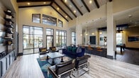 This single-story clubroom in Spring, Texas, is a well-appointed communal space designed for socializing and recreation. The architecture immediately impresses with soaring vaulted ceilings accented by bold, dark-stained exposed wood beams set against light wood-paneled ceiling insets. Recessed lighting and pendant fixtures illuminate the space throughout.

The main lounge area features light wood-look tile flooring and is furnished with a deep navy blue sofa, leather accent chairs with chrome frames, and a dark wood coffee table atop a vibrant abstract area rug. Floor-to-ceiling black-framed windows and glass doors line the exterior walls, flooding the space with natural light and providing views of the surrounding outdoor pool area and landscaping. Built-in shelving with decorative accents anchors one wall, adding both function and style.

Adjacent to the lounge, a wet bar area offers a textured dark tile backsplash, pendant lighting, and counter-height seating with leather barstools. The billiards zone features dark tile flooring, a full-size pool table, a wall-mounted television, and dramatic gold-toned pendant cluster lighting overhead. Quieter seating nooks with black leather chairs and large windows provide additional gathering spots throughout the space.