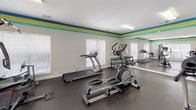Located at 15235 Oneal Rd in Gulfport, Mississippi, this fitness center is a well-equipped, single-floor facility designed to support a full range of exercise routines. The space is immediately defined by its eye-catching décor — bold horizontal stripes in teal, blue, and yellow run along the upper walls, lending the room an energetic and modern feel. The light gray walls and dark rubber flooring provide a clean, functional aesthetic throughout.

The fitness center is outfitted with a comprehensive selection of True-brand cardio equipment, including treadmills, an elliptical trainer, and a recumbent stationary bike, providing options for varied cardiovascular workouts. A multi-function cable strength machine anchors one area of the room, accommodating a wide range of resistance training exercises. A free weight rack with dumbbells and exercise balls adds further versatility to the space.

Multiple windows fitted with white blinds allow natural light to fill the room while maintaining privacy. A large wall mirror expands the sense of space and allows users to monitor their form during workouts. A wall-mounted flat-screen television provides entertainment, and a posted set of fitness center rules ensures a welcoming environment for all users. An adjacent restroom and a vending machine are conveniently accessible through an interior doorway, rounding out the practical amenities of this community fitness facility.