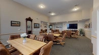 This single-floor community space is thoughtfully designed to foster social interaction and comfort. The layout flows seamlessly across several interconnected gathering areas, each furnished with solid wood dining tables and cushioned rolling chairs, creating a relaxed yet functional atmosphere suited for group activities and shared meals. Multiple seating arrangements accommodate both intimate gatherings and larger group settings, offering flexibility for a variety of uses.

A large flat-screen television is mounted in one of the main gathering areas, accompanied by a media console and decorative indoor plants. The walls throughout are adorned with framed artwork and landscape paintings, adding warmth and visual interest to the neutral-toned interiors. Handrails line the corridors, reflecting a design that prioritizes accessibility and ease of movement.

The fully equipped kitchen features rich dark-stained cabinetry, granite countertops, hardwood-style flooring, and stainless steel appliances including a refrigerator, range, microwave, and dishwasher. The U-shaped layout maximizes workspace and functionality.

Additional highlights include a decorative antique mirror, plush carpeted floors across the common areas, recessed lighting, and natural light filtering in through window treatments. The overall ambiance is warm, inviting, and well-suited for a senior living or assisted living community setting.