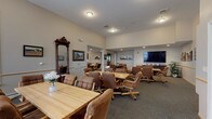 This single-floor community space is thoughtfully designed to foster social interaction and comfort. The layout flows seamlessly across several interconnected gathering areas, each furnished with solid wood dining tables and cushioned rolling chairs, creating a relaxed yet functional atmosphere suited for group activities and shared meals. Multiple seating arrangements accommodate both intimate gatherings and larger group settings, offering flexibility for a variety of uses.

A large flat-screen television is mounted in one of the main gathering areas, accompanied by a media console and decorative indoor plants. The walls throughout are adorned with framed artwork and landscape paintings, adding warmth and visual interest to the neutral-toned interiors. Handrails line the corridors, reflecting a design that prioritizes accessibility and ease of movement.

The fully equipped kitchen features rich dark-stained cabinetry, granite countertops, hardwood-style flooring, and stainless steel appliances including a refrigerator, range, microwave, and dishwasher. The U-shaped layout maximizes workspace and functionality.

Additional highlights include a decorative antique mirror, plush carpeted floors across the common areas, recessed lighting, and natural light filtering in through window treatments. The overall ambiance is warm, inviting, and well-suited for a senior living or assisted living community setting.