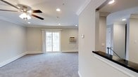 Located in Coram, NY, this single-floor residence offers a clean, well-maintained interior designed for comfortable living. The space features elegant crown molding throughout, adding a refined architectural touch to each room. Recessed lighting illuminates the ceilings consistently, providing bright and even light across the living areas.

The main living space is generously sized, with soft, plush carpeting underfoot and neutral-toned walls that create a calm, welcoming atmosphere. A ceiling fan is centrally positioned overhead, complementing the wall-mounted air conditioning unit for year-round climate control. Sliding glass doors allow natural light to pour in and provide a direct view to the exterior.

A partial wall with a pass-through window connects the living area to an adjacent space, topped with a dark countertop that adds a contrasting design element. A stainless steel appliance is visible through the opening, hinting at a functional kitchen nearby.

The interior corridor features dark hardwood flooring, offering a striking contrast to the carpeted living areas. White panel doors and crisp white baseboards throughout the space contribute to a cohesive, tidy aesthetic. The overall ambiance is bright, neutral, and move-in ready, making this Coram residence a well-appointed and practical single-floor home.